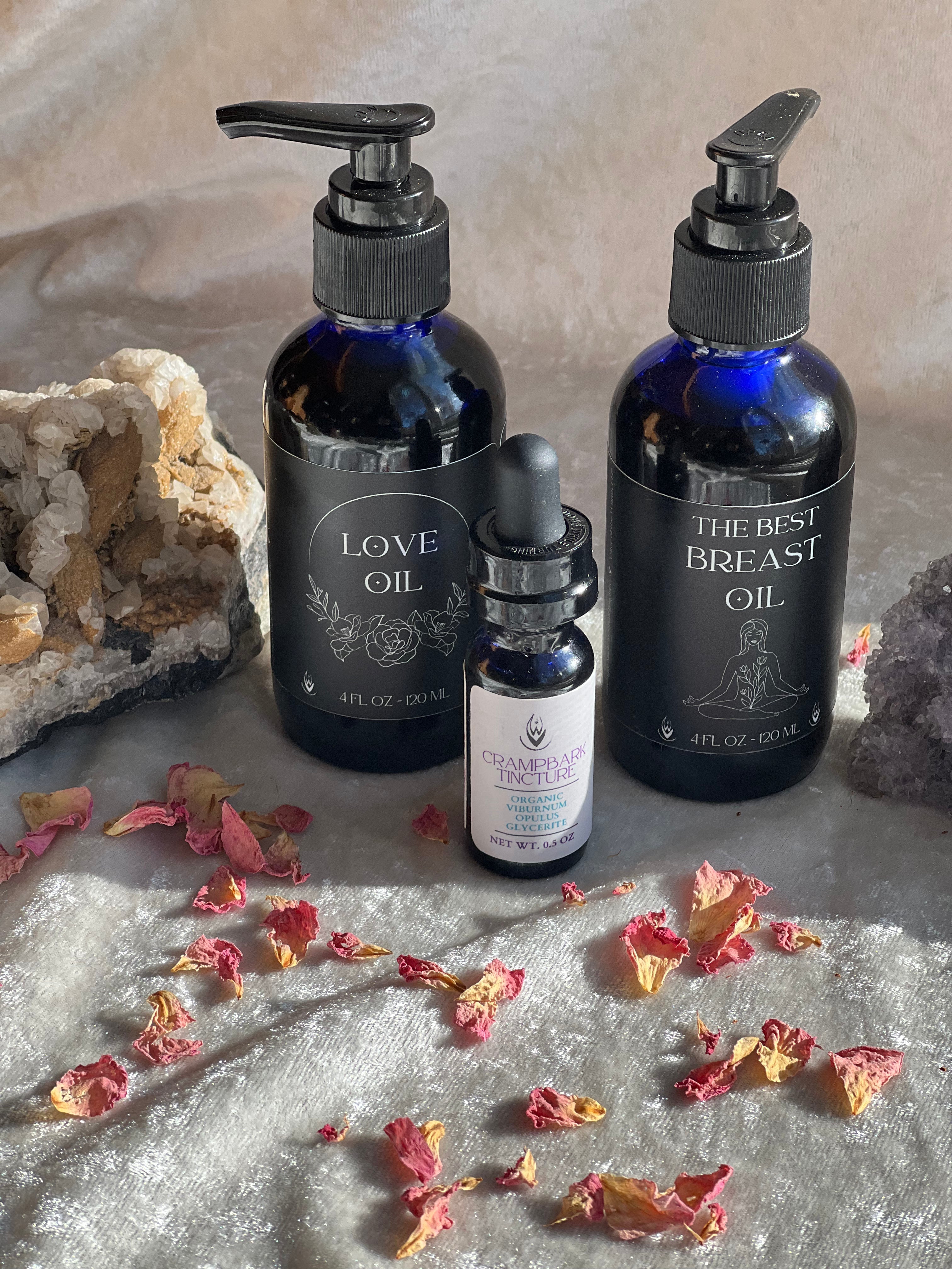 Three bottles of oils labeled 'Love Oil', 'The Best Breast Oil', and a smaller dropper bottle on a textured surface with dried flowers.