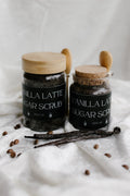 Two jars of vanilla latte sugar scrub with vanilla beans and coffee beans on a white fabric background. velvet bloom beauty and wellness