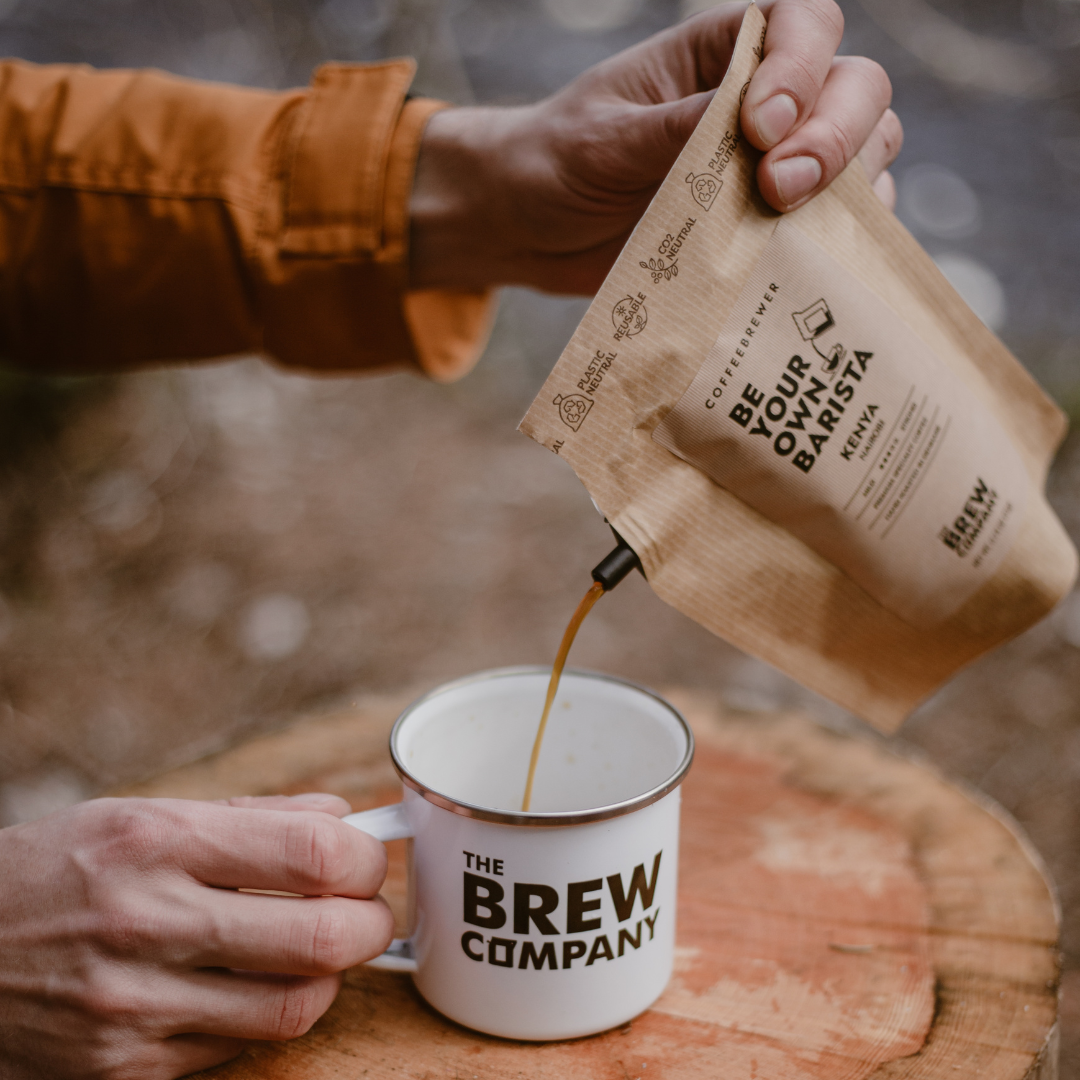 Person pouring coffee from a 'The Brew Company' bag into a white mug on a wooden surface.