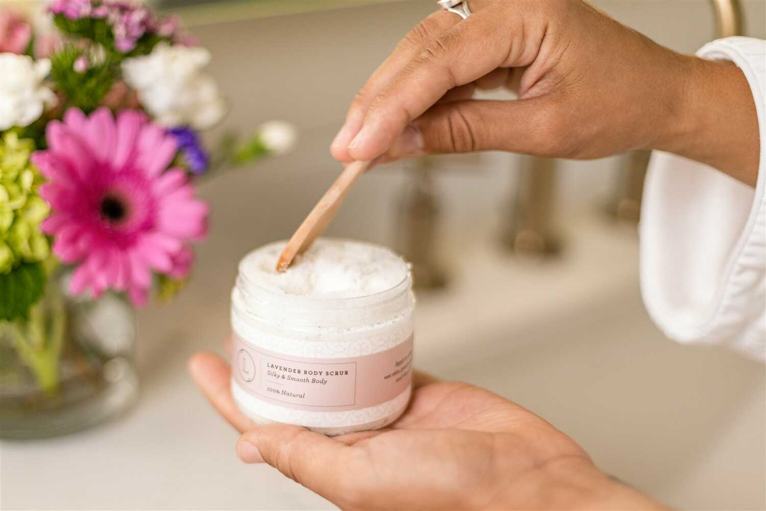 Person using a wooden applicator to scoop out a white cream from a pink jar, with flowers in the background. velvet bloom beauty and wellness